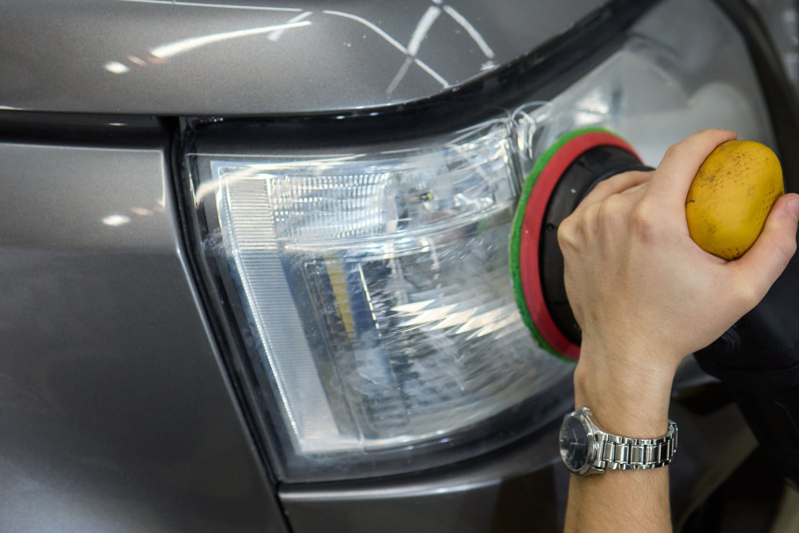 A person is using a machine to polish an automotive headlight, part of the vehicles lighting system, as part of automotive engineering maintenance